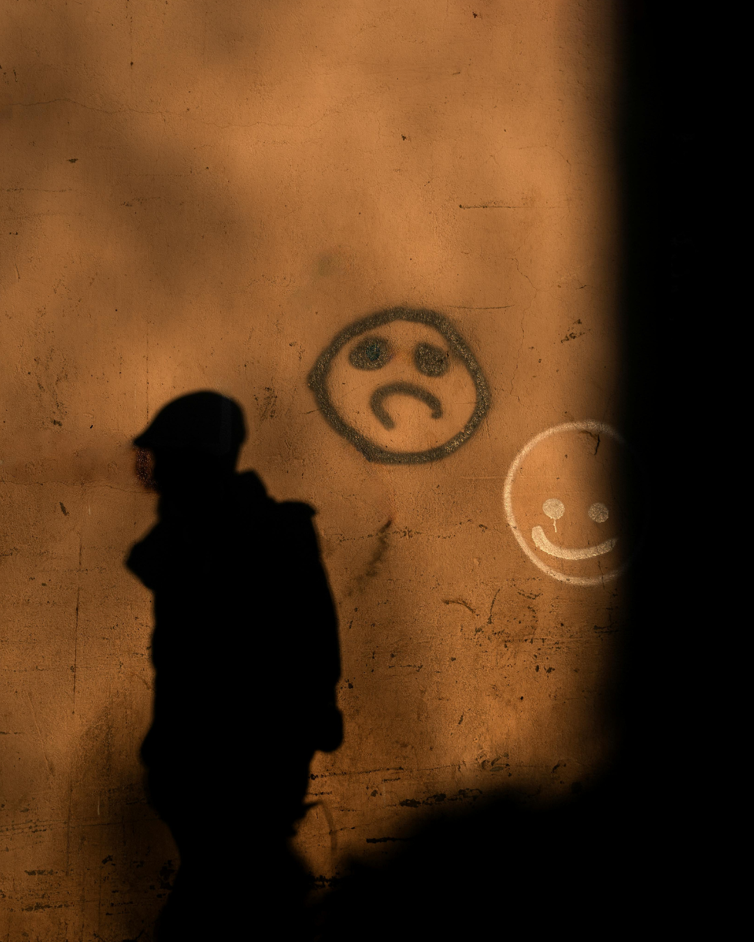Brown, orange wall with a smiley face and a frowning face painted on it. A shadow walking by the wall.